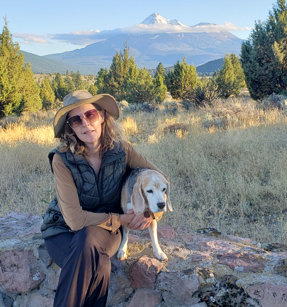 Woman in wide-brim hat kneels with tan dog in sunlit mountain meadow, snow-capped peak behind